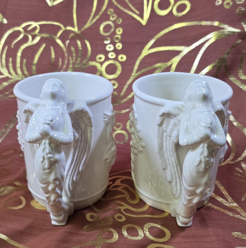 May include: Two white ceramic mugs with angel handles. The handles are sculpted in the form of an angel with outstretched wings and a rose. The mugs have a classic, elegant design, perfect for coffee or tea. The mugs are set against a patterned background.