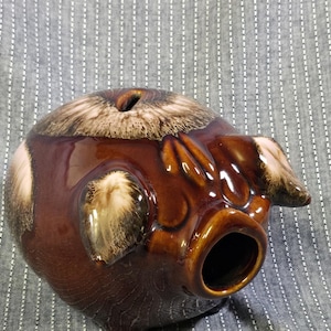 Pode incluir: Um cofrinho de cerâmica marrom com acabamento brilhante. O porco tem um corpo redondo, um focinho e orelhas pequenas. A ranhura da moeda está no topo. O cofrinho é um item decorativo.