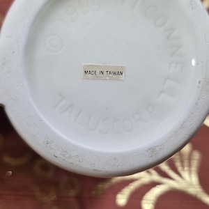 May include: Close-up of the bottom of a white ceramic mug. The text "MADE IN TAIWAN" is visible on a small sticker. The mug's base also includes the text "&copy; 1993 J. CONNELL" and "TALUSCORP".