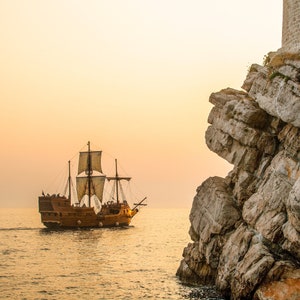 Op de afbeelding: Een houten zeilschip met witte zeilen vaart over het water naar een rotsachtige klif. De lucht is zacht oranje van kleur, wat suggereert dat de zon ondergaat.