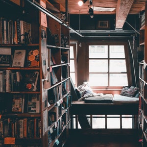 Op de afbeelding: Een houten boekenkast gevuld met boeken, met een vensterbank in de achtergrond. De vensterbank is bedekt met een stoffen bekleding met een patroon en heeft een kussen.