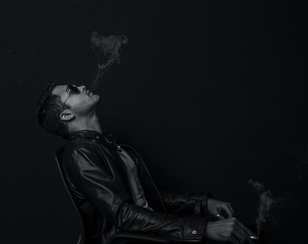 May include: A man in a black leather jacket sits in a chair and smokes a cigarette. He is wearing sunglasses and looking up at the smoke.