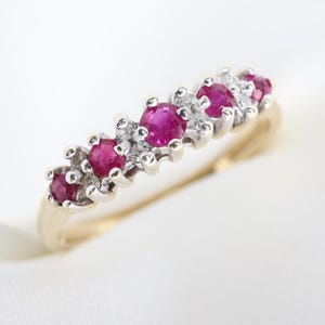 Könnte beinhalten: Ein Goldring mit einer Reihe von fünf runden, leuchtend rosaroten Rubinen, die sich mit kleinen, klaren, funkelnden Akzentsteinen abwechseln, gefasst in einer silberfarbenen Metallfassung auf der Oberseite des Rings.