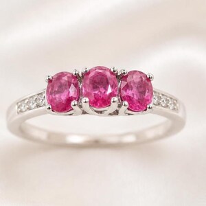 Peut inclure: Une bague en argent ornée de trois pierres précieuses roses vives de forme ovale. La monture est ornée de petites pierres claires. La bague est posée sur un fond blanc et soyeux, soulignant l'élégance du bijou.