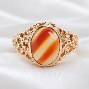 Könnte beinhalten: Ein goldener Ring mit einem ovalen Cabochon-Stein, der diagonale Streifen in Orange und Creme aufweist. Die Ringschiene hat eine texturierte, poröse und unregelmäßige Oberfläche, die die zentrale Steinfassung in einem detaillierten Metallrahmen umgibt.