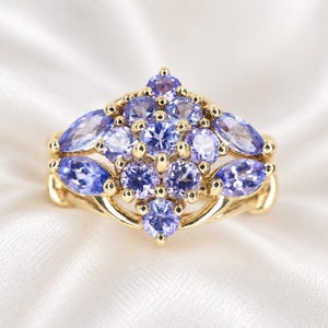 Könnte beinhalten: Ein Goldring mit einer Ansammlung von hellvioletten Edelsteinen im Rund- und Marquiseschliff. Die Steine sind in einem symmetrischen Muster angeordnet, wobei ein zentraler runder Stein von kleineren runden und zwei Marquise-Steinen umgeben ist.