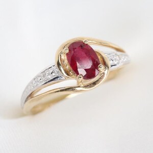 Peut inclure: Une bague ornée d'un rubis rouge profond taille ovale maintenu par quatre griffes. L'anneau est en or jaune et présente un design divisé avec des accents en métal blanc sur les épaules, sertis de petites pierres rondes et transparentes.
