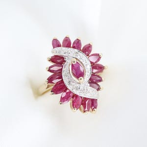 Puede incluir: Anillo de oro con un grupo de rubíes tallados en marquesa y pequeños diamantes. Los rubíes rojo oscuro están dispuestos en un patrón floral alrededor de un rubí central. El anillo tiene un diseño clásico y elegante.