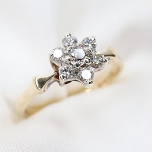Peut inclure: Une bague en or avec un sertissage en forme de fleur composé de sept pierres rondes, claires et facettées. La pierre centrale est entourée de six pierres plus petites, le tout monté sur un support argenté sur un anneau en or jaune poli.