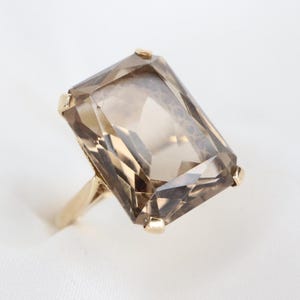 Può includere: Un anello in oro con una grande pietra preziosa di quarzo fumé a taglio rettangolare sfaccettato. La pietra è fissata da quattro piccole griffe in oro su ogni angolo, e la fascia è in metallo dorato lucido che si restringe verso la base.