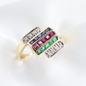 Könnte beinhalten: Ein goldener Ring mit einem rechteckigen Mittelteil mit Reihen aus blauen, roten und grünen Edelsteinen. Der Ring hat Diamantakzente. Der Ring ist vor einem weißen Hintergrund platziert.