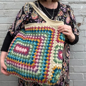 Könnte beinhalten: Eine große, farbenfrohe Häkeltasche mit Granny-Square-Design. Die Tasche hat eine Vielzahl von Farben, darunter Rosa, Blau, Grün und Gelb. Die Griffe und der obere Rand sind beige. Die Tasche wird vor einer grauen Backsteinmauer gehalten.