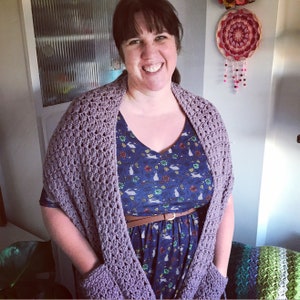 May include: A woman wearing a purple crochet shawl with pockets over a blue dress with a floral and bunny print. The dress has a brown belt.
