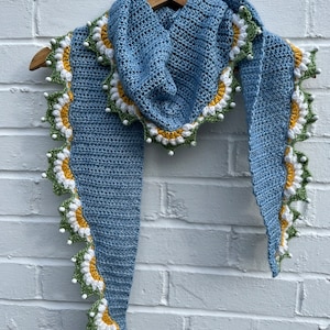 May include: A light blue crocheted scarf with a white, yellow, and green daisy trim. The scarf is hanging on a wooden hanger against a white brick wall.