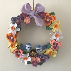 Op de afbeelding: Een gehaakte krans met een paarse strik, met blauwe vogels, gele, witte en paarse bloemen. De krans is versierd met gehaakte bladeren en een nest met eieren.
