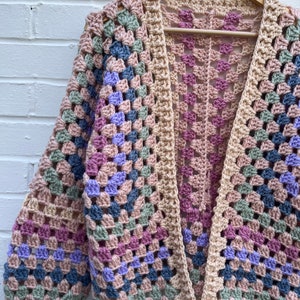 Peut inclure: Un cardigan en crochet avec un motif de granny squares dans des tons de rose, bleu, vert et beige. Le cardigan a un col en V et des manches longues.