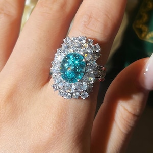 May include: A striking ring with a central oval teal gemstone, encircled by numerous smaller, clear gemstones. The ring's silver band and floral arrangement create a luxurious design. The ring is a beautiful piece of jewellery.