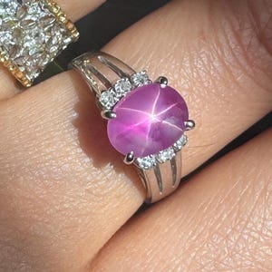 May include: A silver ring featuring a large, oval, pink star ruby gemstone. The ring has a split shank design with small, round, clear gemstones on either side of the ruby. A second ring with a gold band and clear gemstones is also visible.