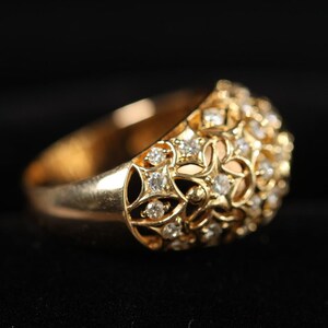 May include: A gold ring with a domed top, encrusted with numerous small diamonds. The ring features an openwork design with floral and geometric patterns. The band is solid gold, and the ring is set against a black background.