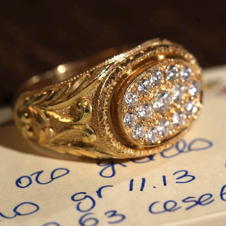 May include: A gold ring with an oval-shaped face set with numerous small, sparkling stones. The ring has intricate floral and scrollwork designs on the band and around the stone setting. The ring is resting on a piece of paper with handwritten text.