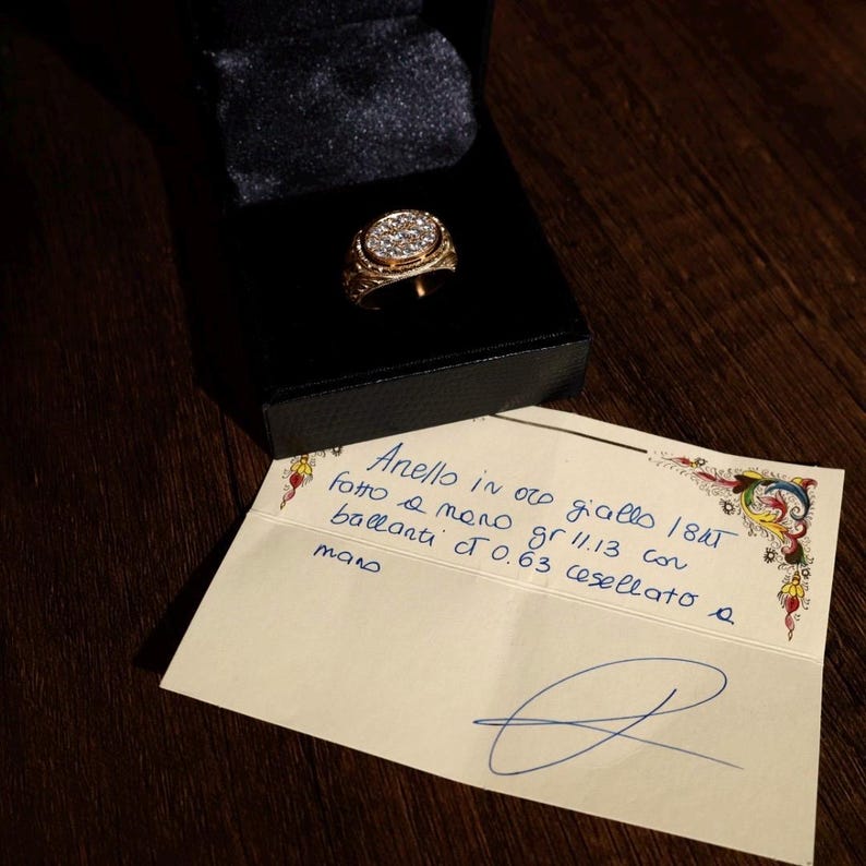 May include: A gold ring with a cluster of small, clear stones set in an oval design. The ring is displayed in a black box. A handwritten note is next to the box.