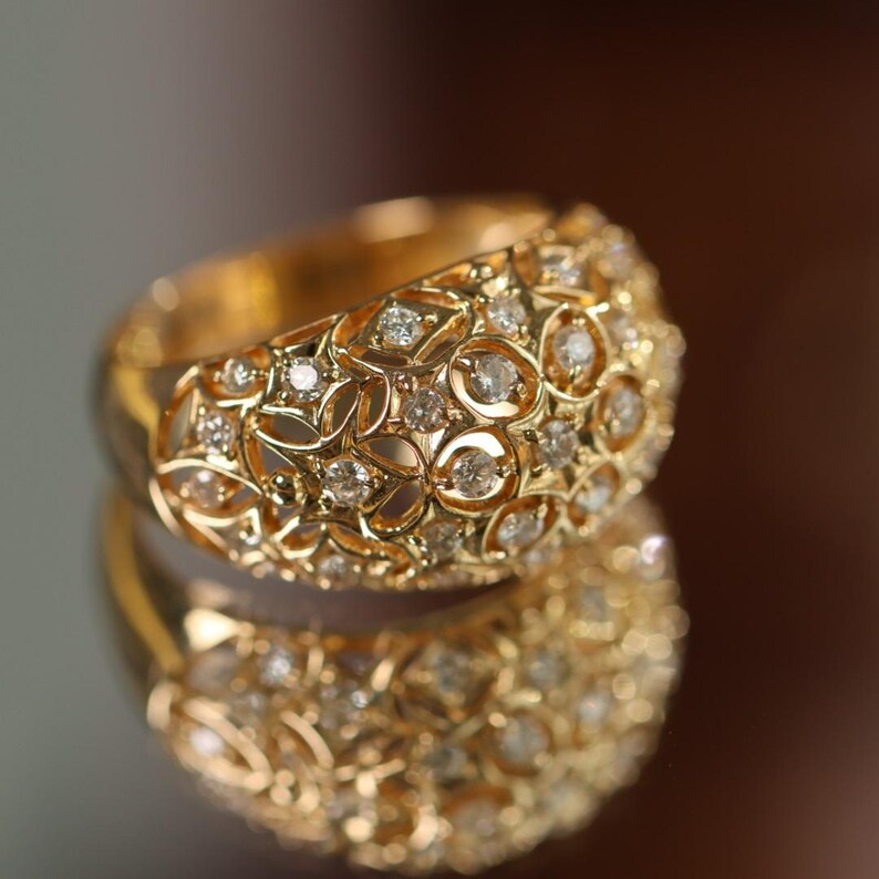 May include: A gold-colored ring with a floral filigree design, encrusted with numerous small, clear gemstones. The ring is reflected in a mirrored surface, enhancing its sparkle and detail.