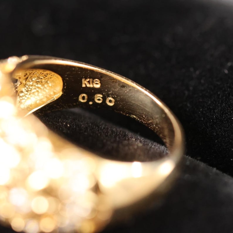 May include: Close-up of a gold ring with the markings "K18" and "0.60" visible. The ring has a textured surface and appears to be made of gold. The background is dark, highlighting the ring's details.