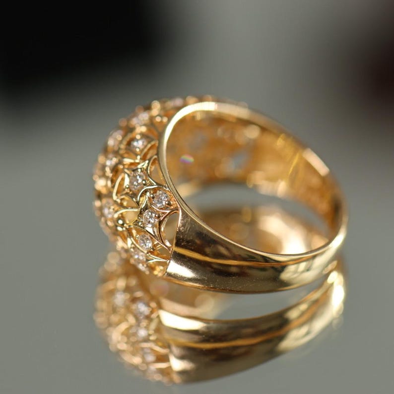 May include: A gold ring with a domed top, encrusted with small, sparkling diamonds. The ring has an intricate, openwork design on the sides, and is reflected in the shiny surface below. The ring is a piece of jewelry.