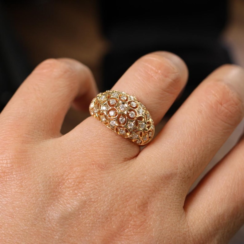 May include: A gold-colored ring with a domed design, encrusted with numerous small, clear gemstones. The ring is worn on a finger, showcasing its intricate, openwork pattern and sparkling details. The ring's surface reflects light, highlighting its luxurious appearance.