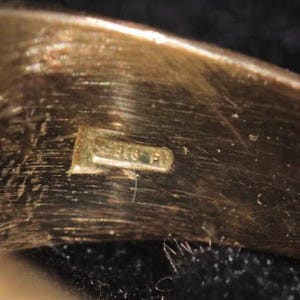 May include: Close-up of a gold-colored ring with a small rectangular hallmark. The hallmark appears to have the numbers "375" and the letters "FI" stamped on it. The ring has a smooth, polished surface.