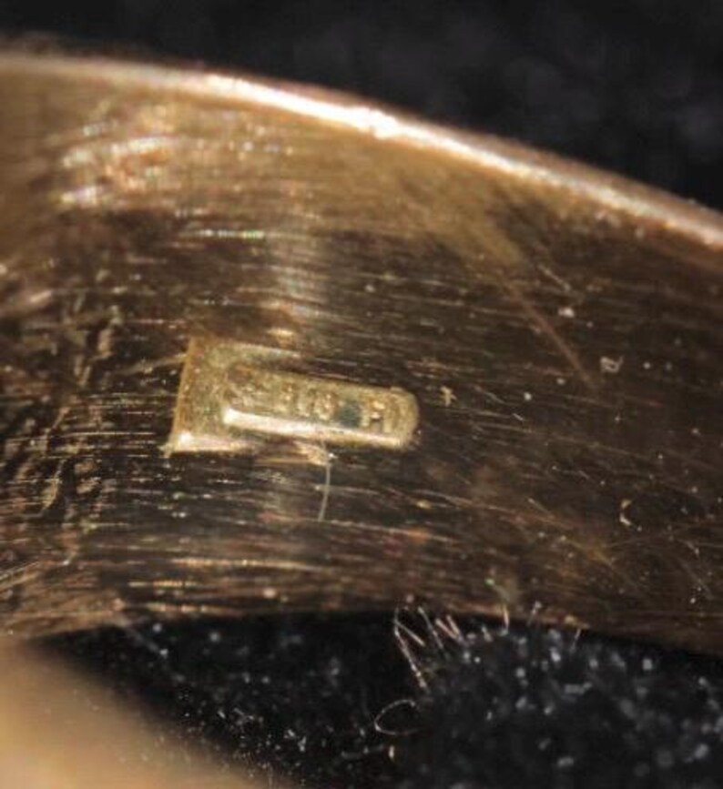 May include: Close-up of a gold-colored ring with a small rectangular hallmark. The hallmark appears to have the numbers "375" and the letters "FI" stamped on it. The ring has a smooth, polished surface.