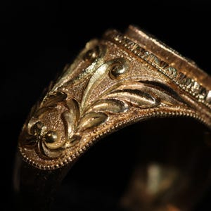 May include: Close-up of a gold ring with intricate floral and leaf engravings. The ring has a textured surface and detailed filigree work. The design features a series of small, beaded accents along the edges, creating a vintage aesthetic.