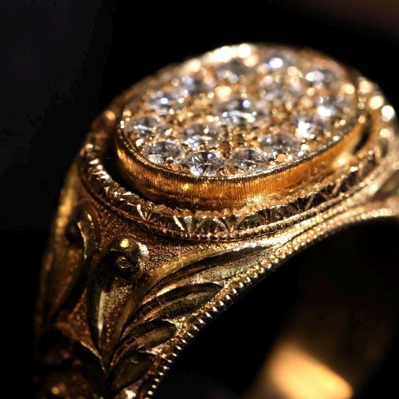 May include: Close-up of a gold ring with an oval-shaped top encrusted with small, clear gemstones. The ring has intricate, engraved floral and geometric patterns. The ring is set against a black background.