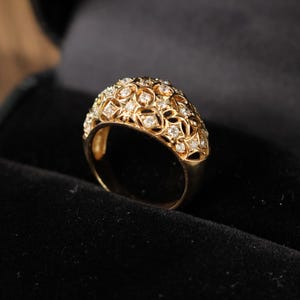 May include: A gold-colored ring with a domed top, encrusted with numerous small, clear gemstones. The ring's design features an open filigree pattern, creating a delicate and ornate appearance. The ring is set against a dark background.