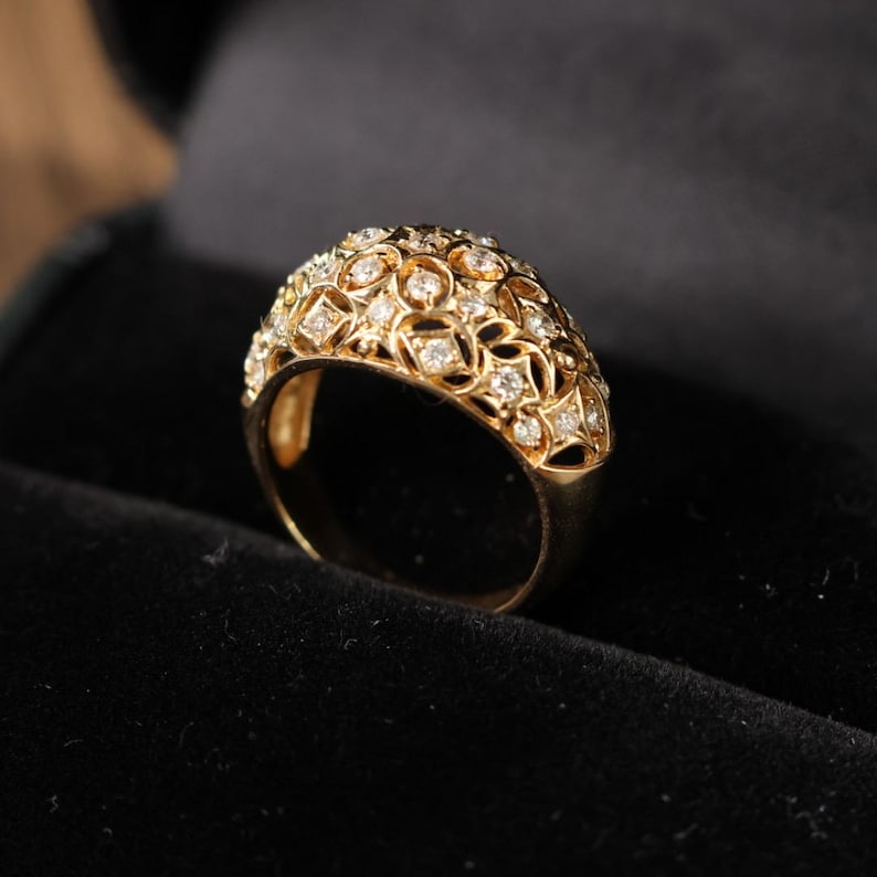 May include: A gold-colored ring with a domed top, encrusted with numerous small, clear gemstones. The ring's design features an open filigree pattern, creating a delicate and ornate appearance. The ring is set against a dark background.
