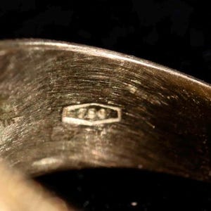 May include: Close-up of a gold ring with a textured surface. The ring has a hallmark stamp with the number "750" inside a rectangular shape. The ring is set against a dark background.