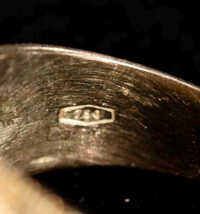 May include: Close-up of a gold ring with a textured surface. The ring has a hallmark stamp with the number "750" inside a rectangular shape. The ring is set against a dark background.