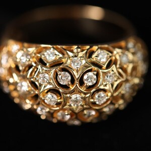 May include: A gold ring with a domed surface, encrusted with numerous small diamonds. The diamonds are set within a detailed, openwork design, creating a sparkling effect. The ring is set against a black background.