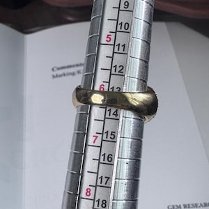 May include: A gold ring is being measured on a ring sizer. The ring is placed on the sizer, which has measurements from 4 to 19. The ring appears to be a size 13. The ring is a simple band style.