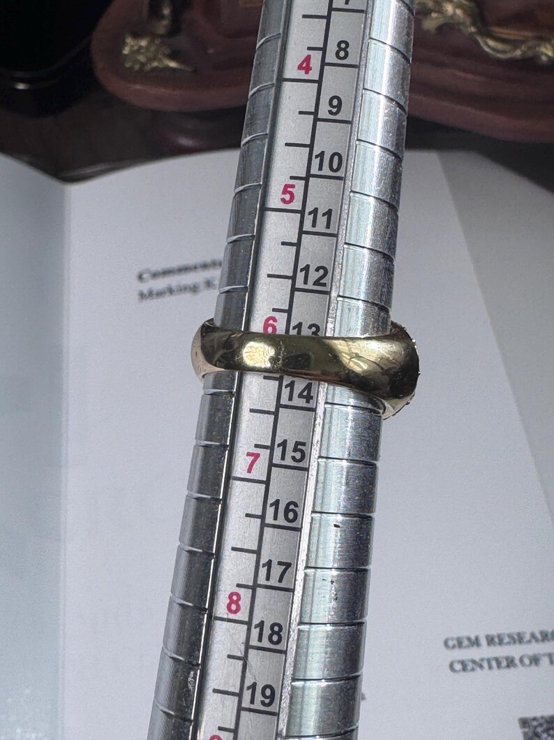 May include: A gold ring is being measured on a ring sizer. The ring is placed on the sizer, which has measurements from 4 to 19. The ring appears to be a size 13. The ring is a simple band style.