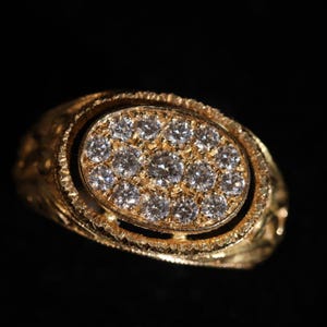 May include: A gold ring with an oval-shaped face, encrusted with numerous small, sparkling diamonds. The ring's band and setting feature intricate, textured details, and the overall design is ornate and luxurious.
