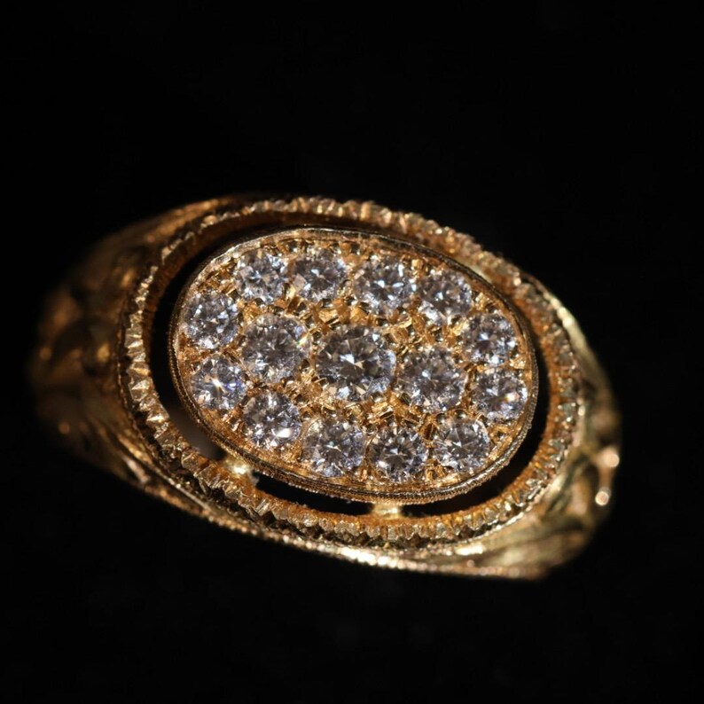 May include: A gold ring with an oval-shaped face, encrusted with numerous small, sparkling diamonds. The ring's band and setting feature intricate, textured details, and the overall design is ornate and luxurious.