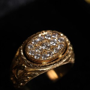 May include: A gold ring with an oval-shaped face, encrusted with numerous small diamonds. The ring has an ornate, engraved design around the band and the diamond setting. The ring is set against a dark background.