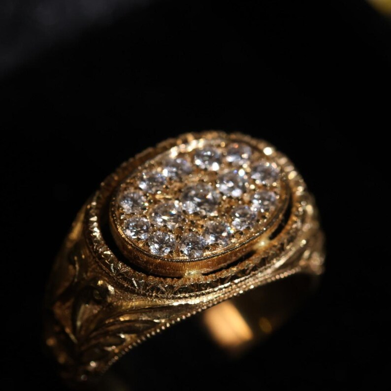 May include: A gold ring with an oval-shaped face, encrusted with numerous small diamonds. The ring has an ornate, engraved design around the band and the diamond setting. The ring is set against a dark background.