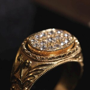 May include: Close-up of a gold ring with a cluster of small, clear gemstones set in an oval shape. The ring has an ornate, engraved design on the band and around the gemstone setting. The ring is a warm gold color.