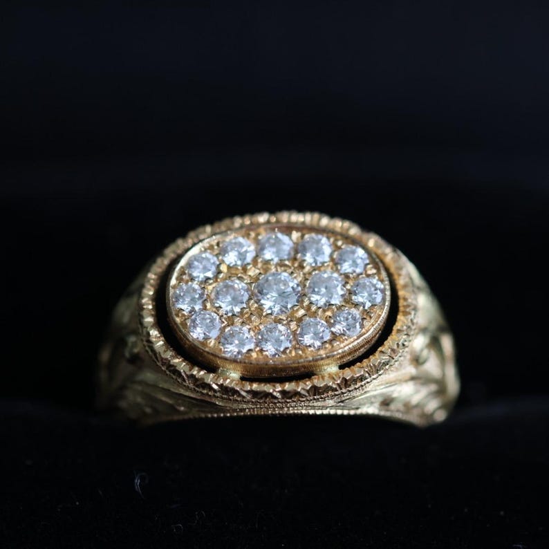 May include: A gold ring with an oval-shaped face set with numerous small, clear gemstones. The ring has an ornate, textured band with a floral design. The gemstones are arranged in a circular pattern.
