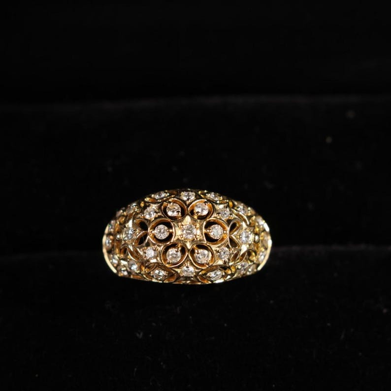 May include: A gold-colored ring with a domed shape, encrusted with numerous small, sparkling diamonds. The ring's surface features an intricate floral pattern, creating a luxurious and ornate appearance. The ring is set against a dark background.