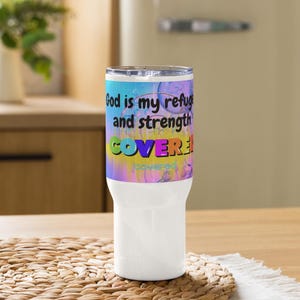 Könnte beinhalten: Ein weißer Becher mit einem farbenfrohen Design. Der Text "God is my refuge and strength COVERED!" ist auf einem Aquarellhintergrund in Blau, Lila und Gelb zu sehen. Der Becher steht auf einer Holzoberfläche.