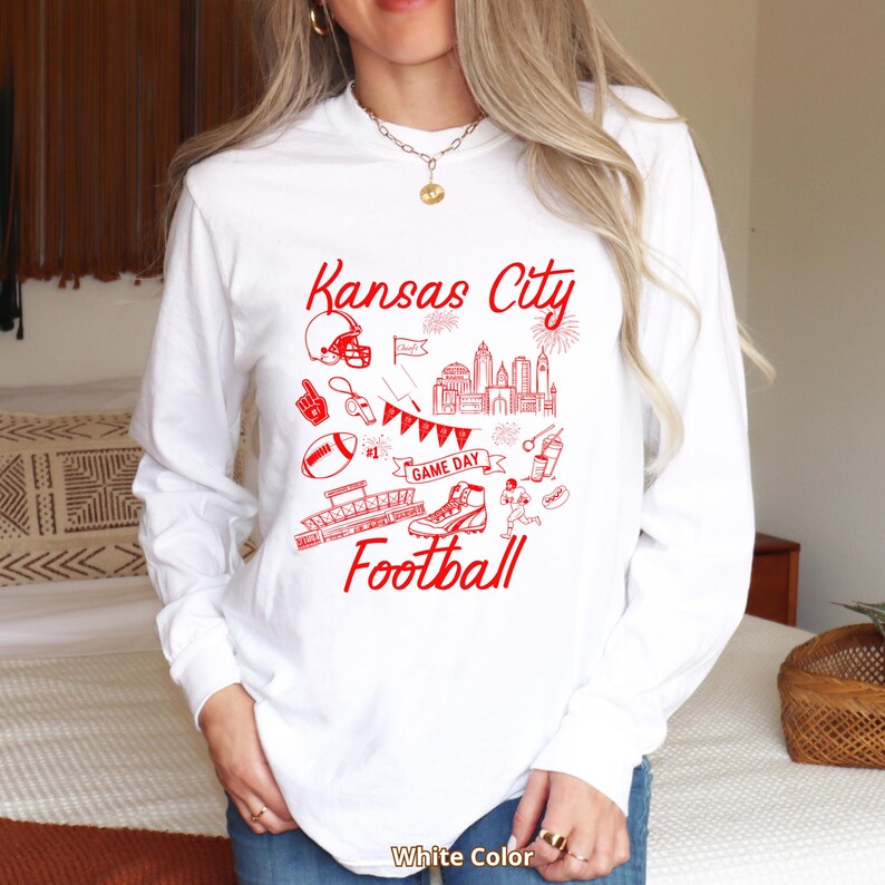 May include: White long-sleeve shirt with a red graphic design. The design includes the text "Kansas City Football" with illustrations of a football helmet, stadium, and other football-related elements. The shirt is a crew neck.