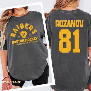 May include: A dark gray t-shirt with the text "RAIDERS BOSTON HOCKEY" in gold lettering. The back of the shirt displays "ROZANOV 81" in gold. The shirt is a Pepper color.